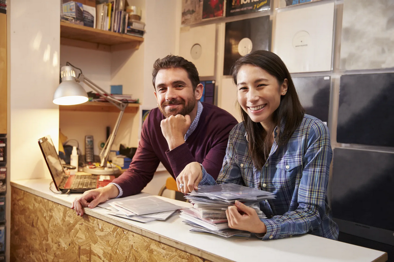 Two people smiling and leaning on a counter, with one holding a stack of vinyl records.