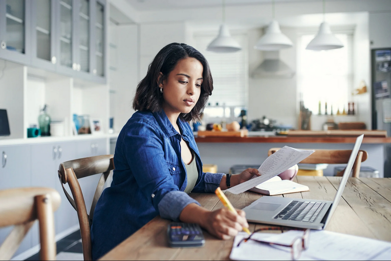 A woman works at a kitchen table with a laptop, papers, and a calculator.