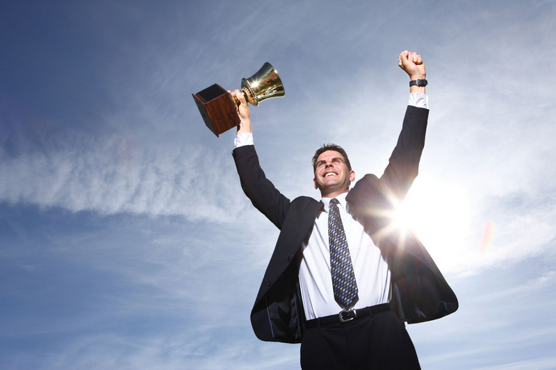 A man in a suit holding a trophy and raising his arms triumphantly against a blue sky background.