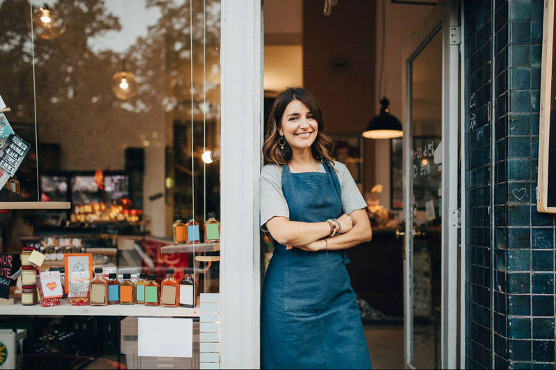 A woman wearing an apron stands smiling at the entrance of a store.