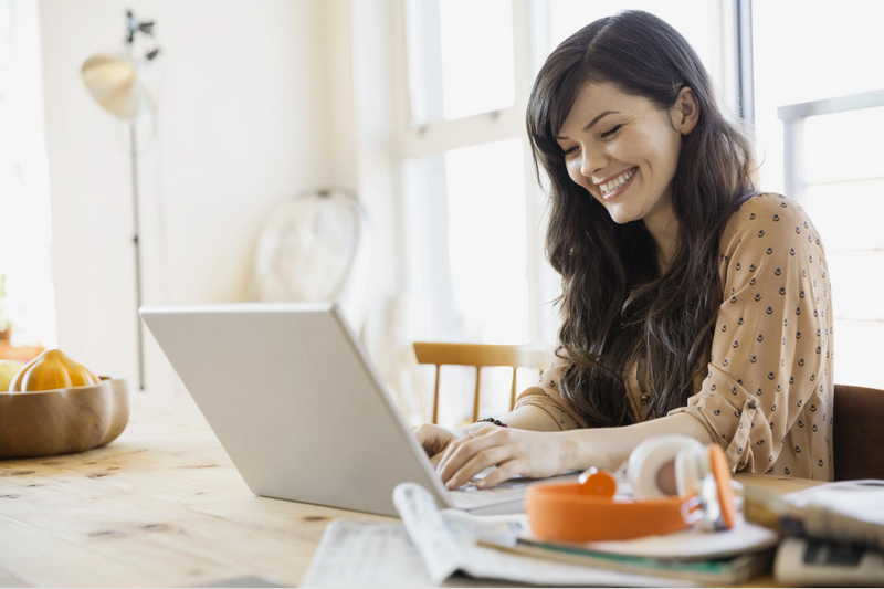 A woman is smiling while using a laptop at a wooden table in a brightly lit room.